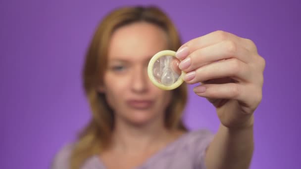 Young beautiful woman, blurred in the background, smiling. in focus is her hand holding an open condom, the concept of safe sex. Protection against AIDS and birth control, color background - Materiał filmowy, wideo