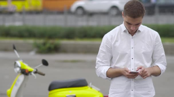 Handsome guy typing a message on the phone. Young guy in white shirt. Handsome boy next to the yellow retro scooter. Busy city street on the background. - Footage, Video