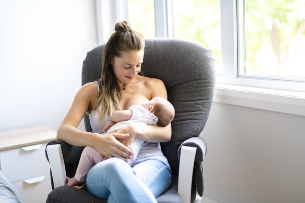 Mother is breastfeeding a baby sitting on a chair close to the window - Foto, imagen