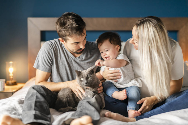 Family of three sit on the bed with 1 years child girl and cat - Valokuva, kuva