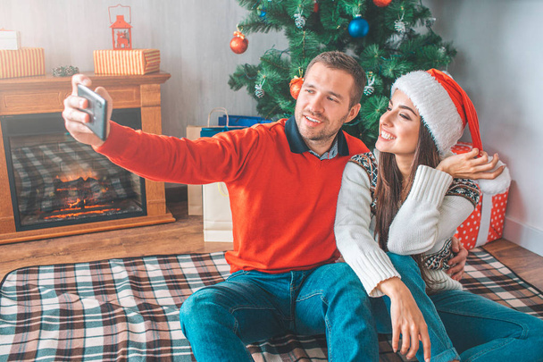 Young man sit on blanket with woman and takes selfie. He embrace her. She poses and smiles. Young woman holds part of her Christams hat. They look happy. - Foto, afbeelding