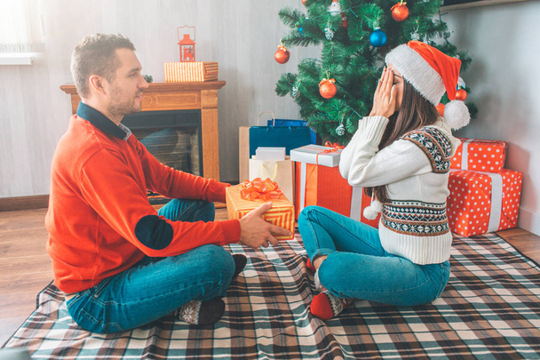 Young man sits in front of woman and looks at her. He holds present. Young woman keeps eyes closed with hands. She wears sweater and Christmas hat. There is tree with presents behind them. - Fotoğraf, Görsel