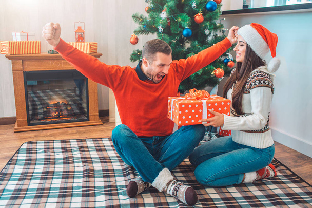 Happy and excited man sits on blanket with fingers crossed and stretches up hands. He joys. Young woman smiles and holds red box with present. She wears Christmas hat. - Photo, Image