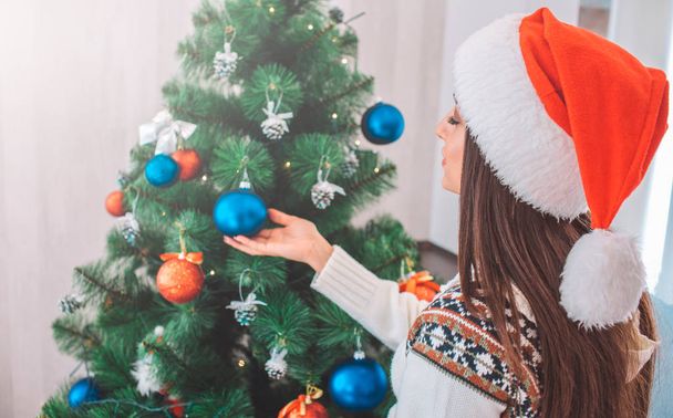 Nice model in red hat stands near Christmas tree and decorates it. She keeps hand under blue toy. Young woman wears Christmas hat. - Фото, зображення