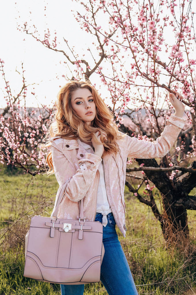 fashion outdoor photo of beautiful sensual girl with blond hair in elegant casual clothes and bag posing in blooming peach garden - Foto, imagen
