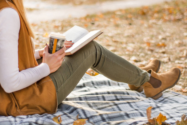 Woman sitting on blanket in park on beautiful autumn day and reading book and holding cup of coffee. Girl wears autumn cloths. Close up, selective focus - 写真・画像