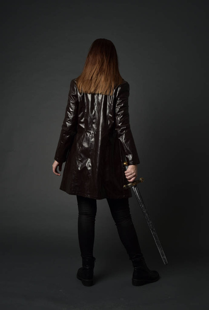 full length portrait of brunette girl wearing  red long leather jack. standing pose, holding a sword on grey studio background. - Fotoğraf, Görsel