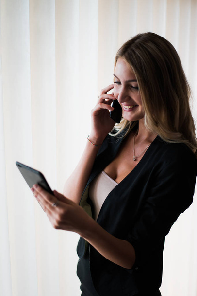 Nuori menestyvä blondi liiketoiminnan nainen puhuu puhelimessa toimistossa ja hymyilee, katselee tablettia
. - Valokuva, kuva