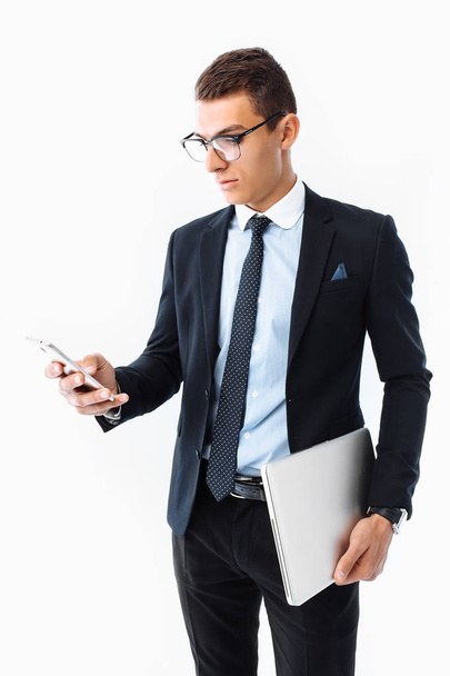 Business man, in a suit and glasses, looking at the phone and holding a laptop in his hands, on a white background - Photo, image