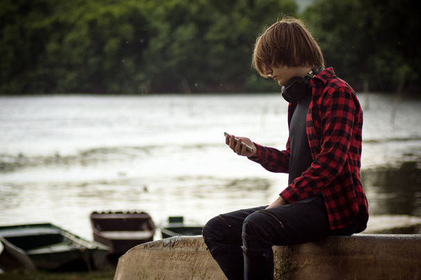 The teenage boy is using phone outdoors at waterside. - Photo, Image
