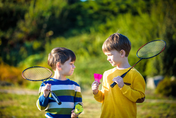 Two active preschool boy playing badminton in outdoor court in summer. Kids play tennis. School sports for children. Racquet and shuttlecock sport for child athlete. Friends happy together warm field. - Photo, image