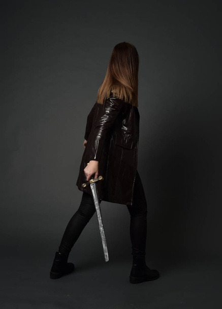 Pełna długość Portret brunetka dziewczyna sobie czerwone skórzane długie jack. Stojący poza, trzymając miecz w studio szary tło. - Zdjęcie, obraz