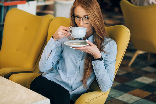 Red-haired girl in a cafe. Is drinking coffee. Checks reports at the computer. - Foto, Bild