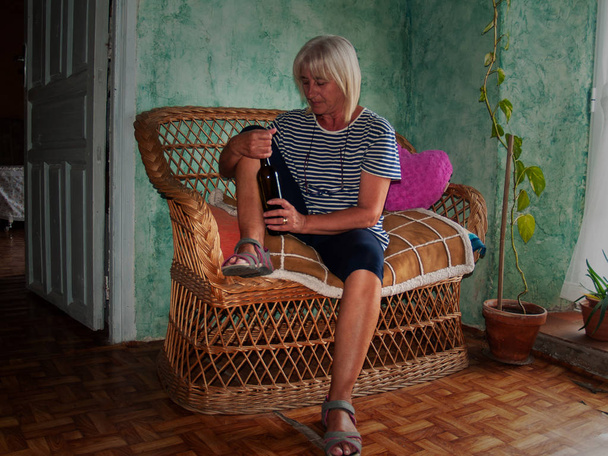Sarhoş bir kadın kıdemli bir kanepe oturma onun elinde bir şişe şarap ile - Fotoğraf, Görsel