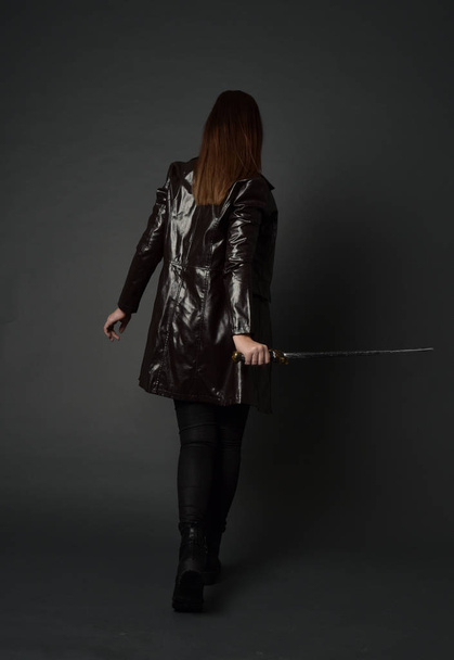 Pełna długość Portret brunetka dziewczyna sobie czerwone skórzane długie jack. Stojący poza, trzymając miecz w studio szary tło. - Zdjęcie, obraz