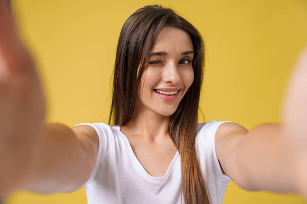 Ευχάριστη ελκυστικό κορίτσι κάνει selfie στο στούντιο και γελώντας. Όμορφος νεαρή γυναίκα με καστανά μαλλιά λαμβάνοντας εικόνα για τον εαυτό της σε φωτεινό κίτρινο φόντο. - Φωτογραφία, εικόνα