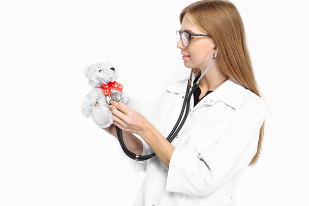 hermosa aprendiz médico con gafas, chica, escuchar a un oso de peluche con un estetoscopio, aislado sobre un fondo blanco
 - Foto, imagen