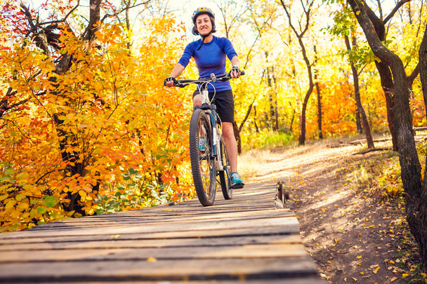 The girl drives on a wooden springboard. Woman riding her bike in the forest. Preparing to jump. Extreme hobby. Cyclist in the autumn park. - Photo, Image