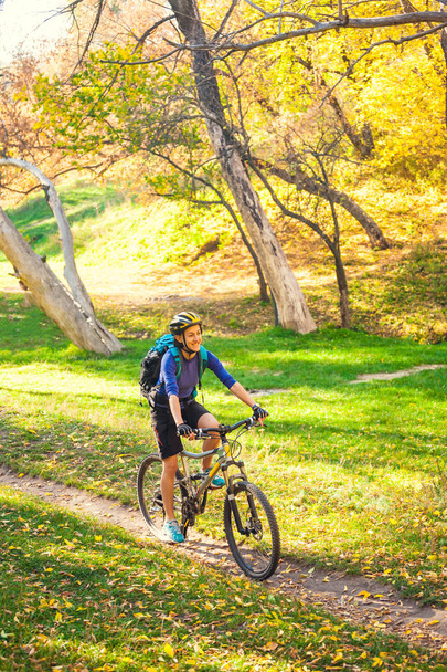 Το κορίτσι με ένα σακίδιο βόλτες με ένα ποδήλατο στο πάρκο φθινόπωρο. Λεπτή γυναίκα τρένα στη φύση. Σπορ στο δάσος. Τουριστικές βόλτες με ένα ίχνος βρωμιά. Ταξιδεύοντας με το ποδήλατο. - Φωτογραφία, εικόνα