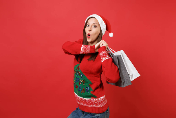 Amazed young Santa girl keeping mouth wide open looking surprised holding packages bags with purchases after shopping isolated on red background. Happy New Year 2019 celebration holiday party concept - Фото, изображение