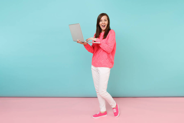 Full length portrait of woman in knitted sweater, white pants using laptop pc computer isolated on bright pink blue pastel wall background in studio. Fashion lifestyle concept. Mock up copy space - Фото, зображення