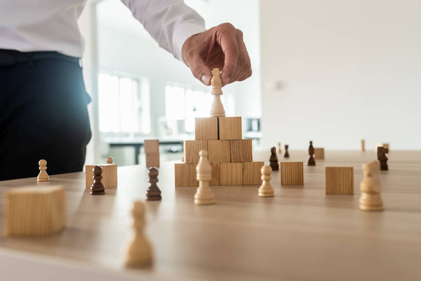 Business hierarchy concept with businessman placing chess figure of king on top of wooden stacked wooden blocks and other figures spread on office desk. - Фото, зображення