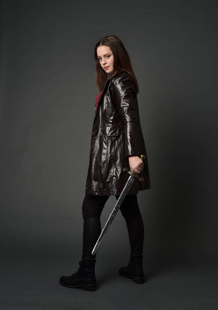 Ganzkörperporträt eines brünetten Mädchens, das einen roten langen Lederjacke trägt. stehend in Pose, mit einem Schwert auf grauem Studiohintergrund. - Foto, Bild