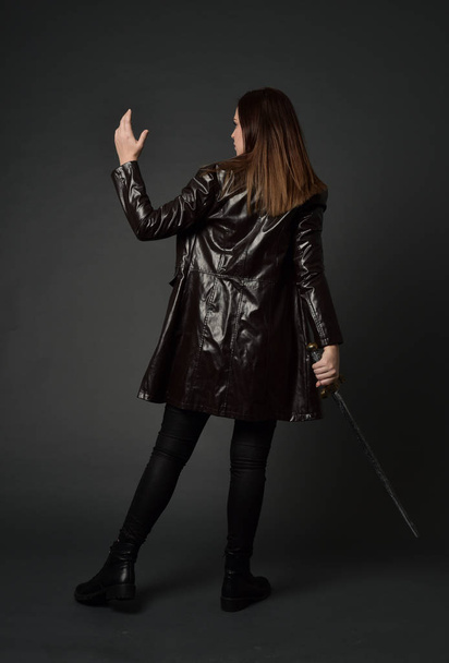 full length portrait of brunette girl wearing  red long leather jack. standing pose, holding a sword on grey studio background. - Photo, Image