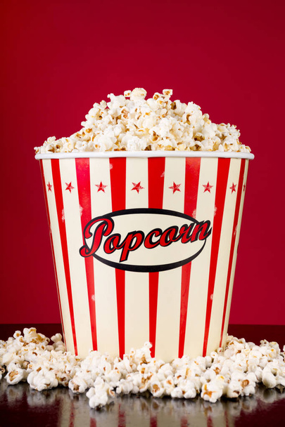 Retro-Box mit Popcorn und verschüttet auf schwarzem reflektierendem Schreibtisch und rotem Hintergrund. Film- und Kino-Food-Konzept. Nahaufnahme - Foto, Bild