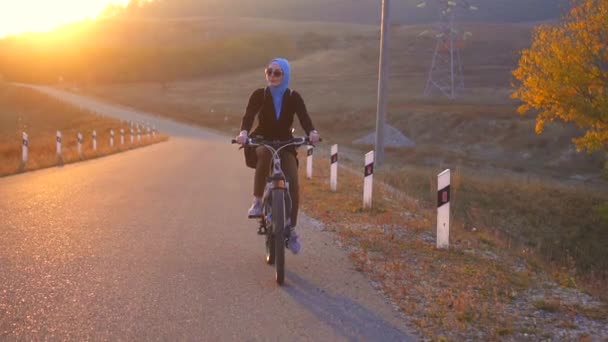 Muzułmanka w hidżab z plecaka, jazda na rowerze na drodze zachodzącego słońca, wolno mo - Materiał filmowy, wideo