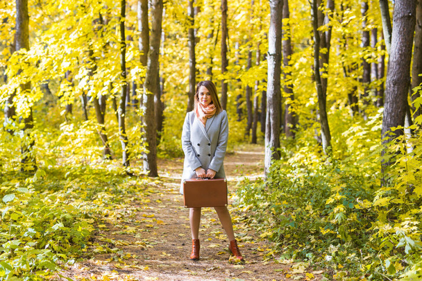 fashion, season and people concept - happy young woman is going on a trip with retro suitcase on a background of autumn foliage - Photo, Image
