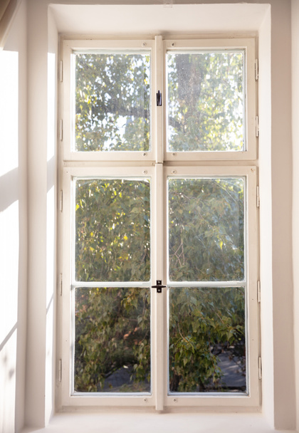 Λευκό παλιό ντεμοντέ ξύλινο παράθυρο στον λευκό τοίχο, πράσινα δέντρα έξω από το - Φωτογραφία, εικόνα