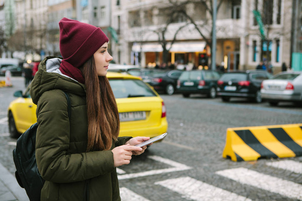 Όμορφα τουριστικά κορίτσι με ένα δισκίο σε έναν δρόμο στην Πράγα καλεί ένα ταξί ή εξετάζει ένα χάρτη ή χρησιμοποιεί το διαδίκτυο ή μια εφαρμογή για κινητά. - Φωτογραφία, εικόνα