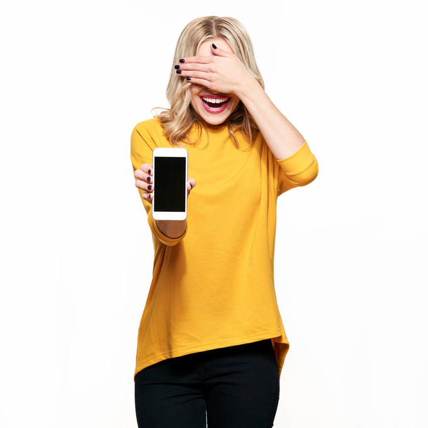 Gorgeous laughing woman showing blank screen mobile phone over white background, covering her eyes with hand. Very excited, cheering emotion. - Foto, immagini