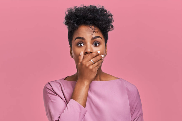 Horizontal shot of surprised African American woman covers mouth with both hands, tries to be speechless, looks with shocked expression, notices something strange, isolated over pink blank wall - Foto, imagen