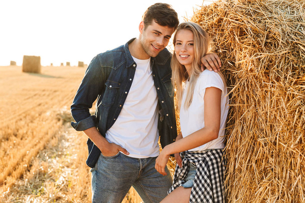 Image of smiling couple man and woman walking on golden field and standing near big haystack - Фото, изображение