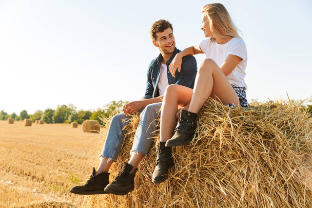 Smiling young couple sitting together on a haystack at the field, talking - Zdjęcie, obraz