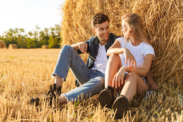 Photo of young couple man and woman sitting under big haystack in golden field during sunny day - Fotó, kép