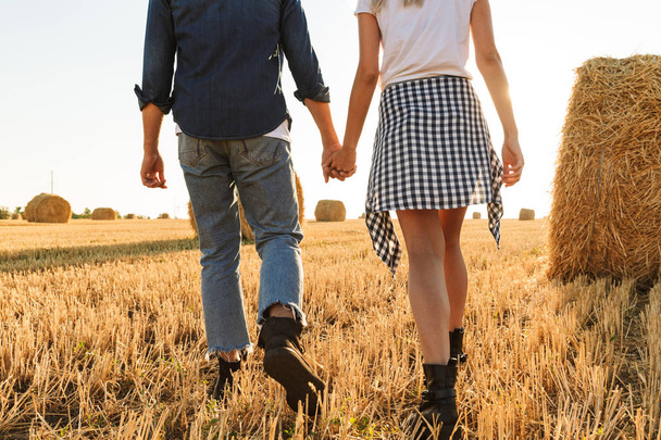 Cropped photo of young guy and girl walking through golden field with bunch of haystacks during sunny day - Foto, Imagen