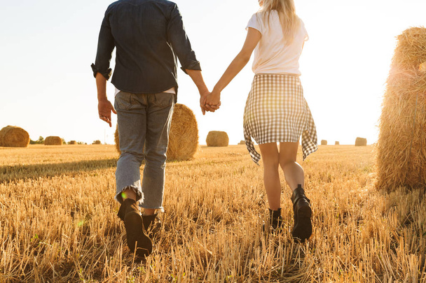 Cropped photo of guy and girl walking hand by hand through golden field with bunch of haystacks during sunny day - Фото, зображення