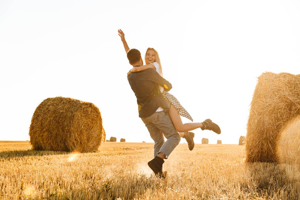 Photo of happy couple man and woman having fun while walking through golden field with bunch of haystacks during sunny day - Foto, Imagem