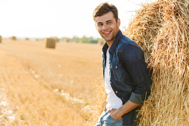 Image of joyful man 30s walking through golden field and standing near big haystack during sunny day - Foto, Bild