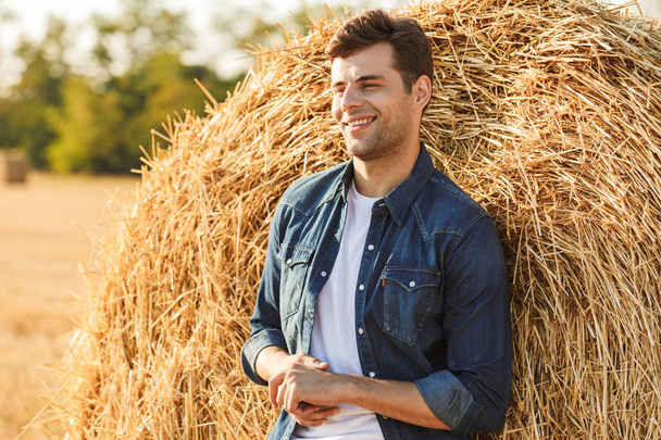 Image of smiling man 30s walking through golden field and standing near big haystack during sunny day - Фото, зображення