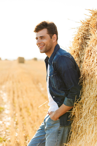 Full length photo of stylish man 30s walking through golden field and standing near big haystack during sunny day - Foto, Imagen
