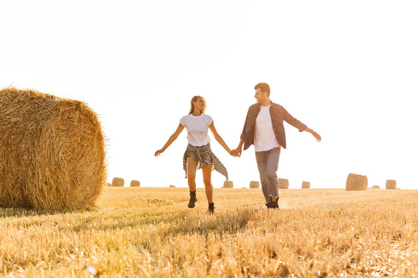 Romantic photo of young man and woman running through golden field with bunch of haystacks during sunny day - Zdjęcie, obraz