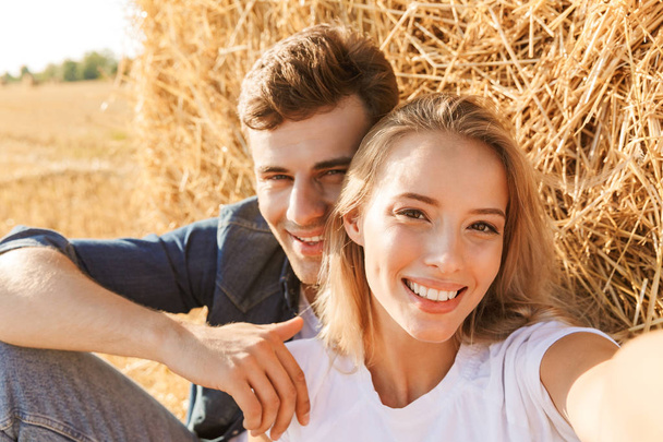 Photo of beautiful couple man and woman taking selfie while sitting under big haystack in golden field during sunny day - Fotó, kép