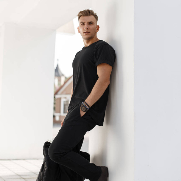 Joven modelo de moda hombre con bolso en camisa negra y pantalón con bolsa negra se para cerca de la pared blanca
 - Foto, Imagen