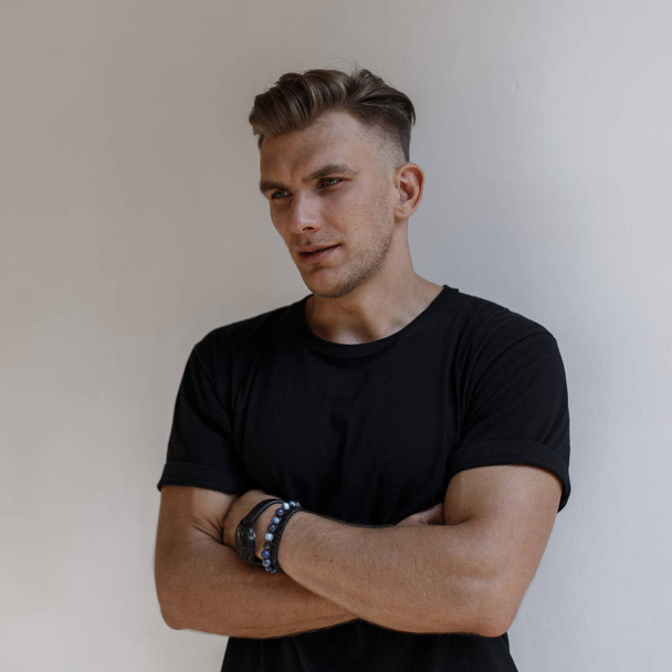 Stylish handsome American man with hairstyle in fashionable black t-shirt posing near a white wall - Photo, Image