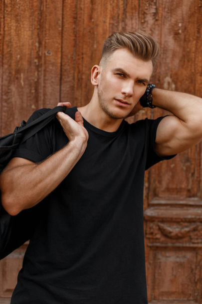 Fashionable handsome man with a hairstyle in a black T-shirt with a black bag stands near the vintage wooden door - Фото, изображение