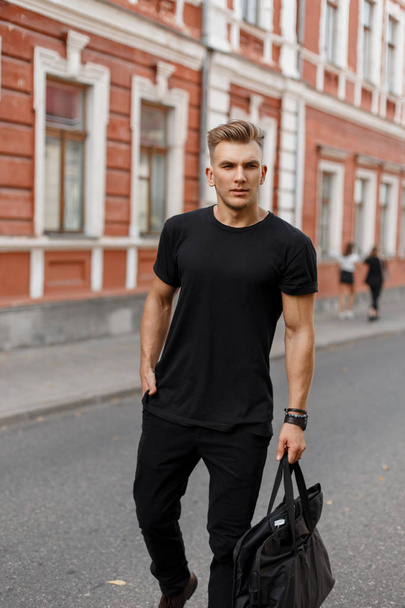 Μοντέρνα όμορφος νεαρός κομψό μοντέλο άνθρωπος με χτένισμα σε μαύρο κομψό ρούχα με μια μαύρη τσάντα που περπατά στο δρόμο στην πόλη - Φωτογραφία, εικόνα